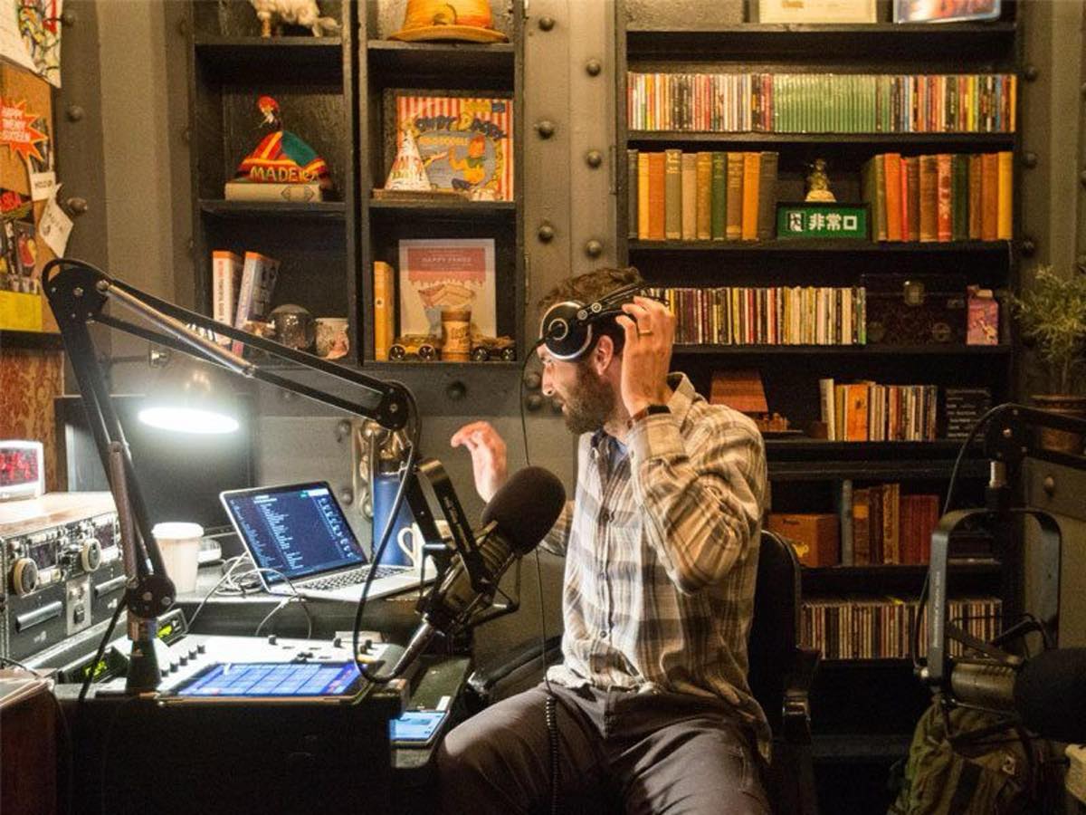Chris Bunting in a radio studio, pulling on headphones beside a boom microphone and laptop, with shelves of CDs and books behind him.